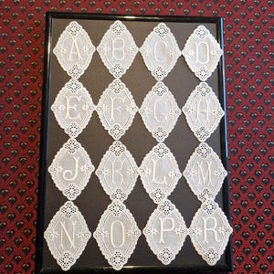 Puede incluir: Una colección enmarcada de letras del alfabeto bordadas en blanco, cada una dentro de un marco en forma de diamante. Se ven las letras A, B, C, D, E, F, G, H, J, K, L, M, N, O, P y R. El bordado presenta delicados patrones florales.