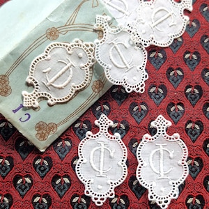 May include: Several white embroidered lace appliqués featuring the letter C in a decorative script font. Each piece has a scalloped edge and a pointed top. The appliqués are displayed on a light green paper packet labeled Art. 7154.