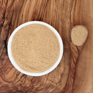 May include: A white bowl filled with light brown nutritional yeast powder, with a small amount of the powder in a wooden spoon beside it.