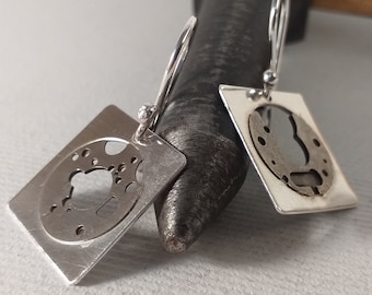 Peut inclure: Une paire de boucles d'oreilles en argent avec des pendentifs de forme carrée. Chaque pendentif a une découpe circulaire avec un motif détaillé.
