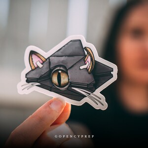May include: A gray and black cat-shaped sticker with a large yellow eye and pink ears. The sticker is on a white background and is being held by a hand.