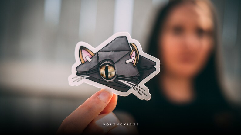 May include: A gray and black cat-shaped sticker with a large yellow eye and pink ears. The sticker is on a white background and is being held by a hand.
