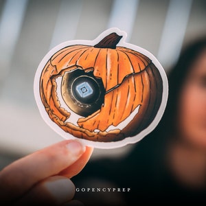 May include: A sticker of a cracked orange pumpkin with a blue and white eye inside. The sticker is held in a hand with the text "GOPENCYPREP" visible at the bottom.