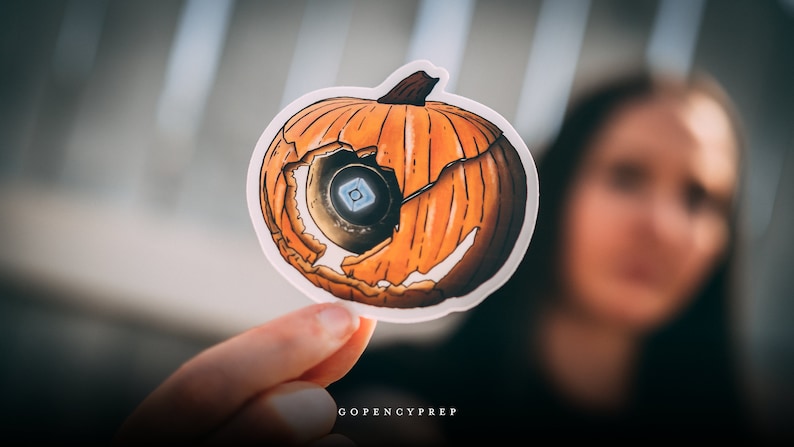 May include: A sticker of a cracked orange pumpkin with a blue and white eye inside. The sticker is held in a hand with the text "GOPENCYPREP" visible at the bottom.