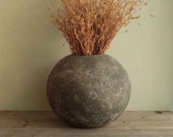 May include: A round, dark gray ceramic vase with a textured surface, filled with dried brown grasses.