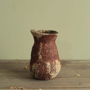 May include: A small, textured vase with a rustic appearance. The vase is primarily a deep reddish-brown color, with patches of off-white and black speckling. The opening is slightly flared, and the vase sits on a wooden surface.