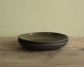 Może przedstawiać: Dwie ułożone w stos płytkie miski ceramiczne z ciemną matową powierzchnią zewnętrzną i gładkim, nakrapianym oliwkowozielonym szkliwem wewnątrz. Miski mają organiczny, lekko nieregularny okrągły kształt z niskimi, zakrzywionymi bokami.