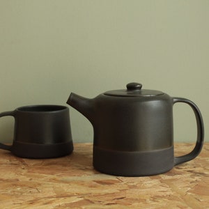 May include: A black ceramic teapot and mug set. The teapot has a rounded body and a spout on the side. The mug has a handle and a rounded body. Both pieces have a dark glaze.