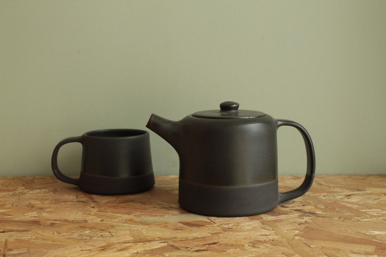 May include: A black ceramic teapot and mug set. The teapot has a rounded body and a spout on the side. The mug has a handle and a rounded body. Both pieces have a dark glaze.