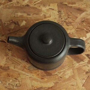 May include: A black ceramic teapot with a lid and a handle. The teapot is sitting on a wooden surface.