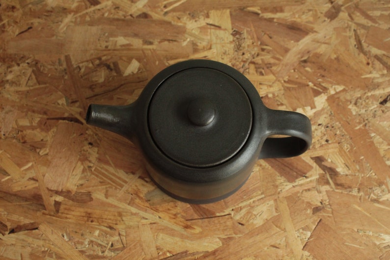 May include: A black ceramic teapot with a lid and a handle. The teapot is sitting on a wooden surface.