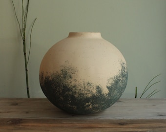 May include: A round ceramic vase with a speckled beige glaze and a dark green ombre design. The vase is sitting on a wooden surface.