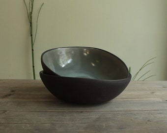 May include: Two ceramic bowls, one nested inside the other. The top bowl is a light grey colour with a glossy finish. The bottom bowl is a dark grey colour with a matte finish.
