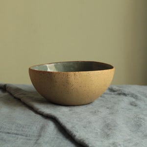 May include: A speckled beige ceramic bowl with a light blue glaze inside. The bowl is sitting on a grey fabric surface.