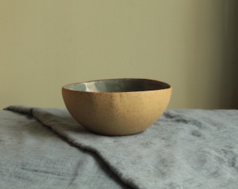 May include: A speckled beige ceramic bowl with a light blue glaze inside. The bowl is sitting on a grey fabric surface.