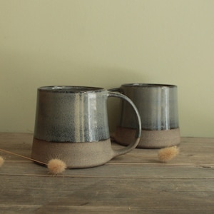 May include: Two ceramic mugs with a light grey glaze and a darker grey rim. The mugs have a speckled, textured finish and a rounded bottom.