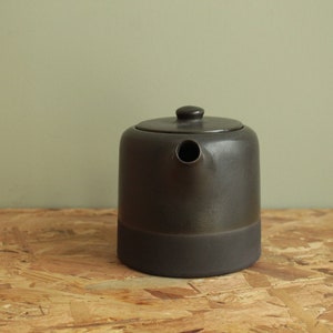 May include: A dark gray ceramic teapot with a wide base and a spout. The teapot has a matte finish and a slightly textured surface.