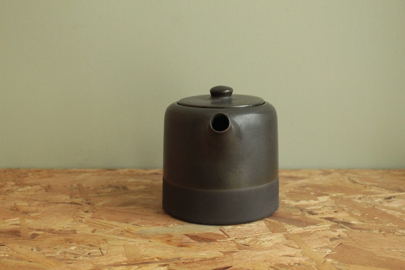 May include: A dark gray ceramic teapot with a wide base and a spout. The teapot has a matte finish and a slightly textured surface.