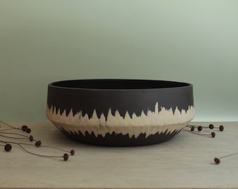 May include: A large, black ceramic bowl with a white, wavy design around the rim. The bowl is sitting on a light brown wooden surface.