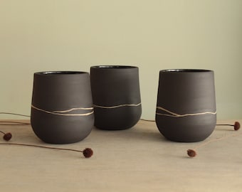 May include: Three black ceramic mugs with a thin gold line design. The mugs are sitting on a wooden surface.