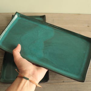 Könnte beinhalten: Ein rechteckiges Keramiktablett mit einer leuchtend türkisfarbenen Glasur und dunklen, sichtbaren Rändern. Die Oberfläche hat ein glattes, glänzendes Finish mit subtilen Farbvariationen, die einen tiefen blau-grünen Ton auf der Basis zeigen.