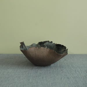 May include: A handmade ceramic bowl with an unusual, natural form. The bowl has a textured, brown outer surface and a dark, speckled inner surface. The rim has a jagged, irregular edge, enhancing its artistic quality. The bowl is likely for decorative use.