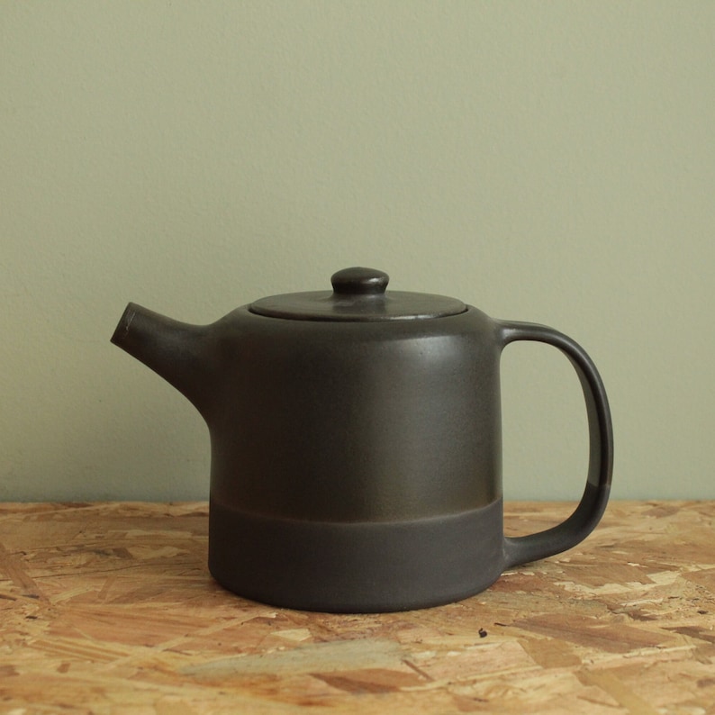 May include: A dark gray ceramic teapot with a wide spout and a handle. The teapot has a matte finish and a slightly rounded shape.