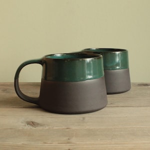 May include: Two ceramic mugs with a dark green glaze on the top half and a black glaze on the bottom half. The mugs are sitting on a wooden surface.