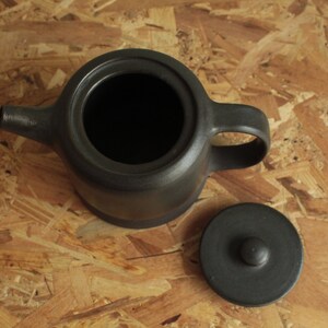 May include: A black ceramic teapot with a lid and a handle. The teapot is sitting on a wooden surface.