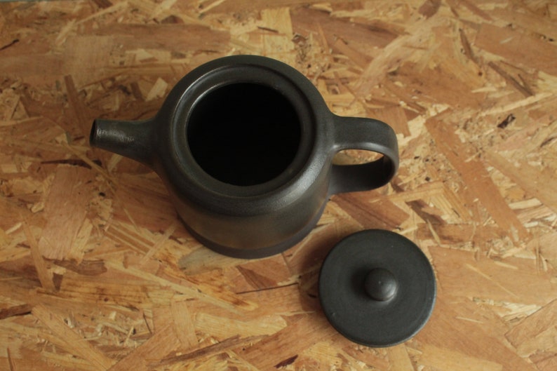 May include: A black ceramic teapot with a lid and a handle. The teapot is sitting on a wooden surface.