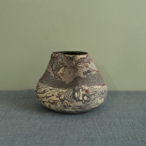 May include: A handcrafted ceramic vase with a unique marbled design. The vase features a bulbous shape with a wide base and a narrower opening. The color palette includes shades of gray, cream, and red, creating a striking visual texture. The vase is a decorative item.