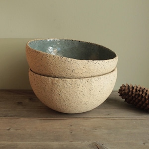 May include: Two handmade ceramic bowls stacked. The bowls have a speckled, beige exterior and a blue-grey interior. A pine cone rests on a wooden surface beside the bowls.