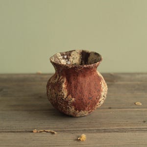 May include: A small, handcrafted ceramic vase with a rustic, textured appearance. The vase features a rounded body and a slightly flared rim. The exterior is primarily a warm, earthy red-brown color, with lighter, mottled patches. The vase sits on a wooden surface.