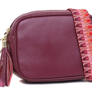 May include: A burgundy crossbody bag with a textured finish. It features a colourful woven strap in shades of red, pink, and orange. The bag has gold-tone hardware and a matching tassel.