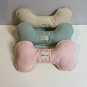 May include: Three dog bone-shaped toys in beige, teal, and pink. Each toy has a white label that reads "SIMPLY FAB". The toys are made of a soft fabric and are stacked on top of each other.