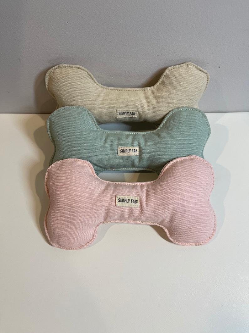 May include: Three dog bone-shaped toys in beige, teal, and pink. Each toy has a white label that reads "SIMPLY FAB". The toys are made of a soft fabric and are stacked on top of each other.