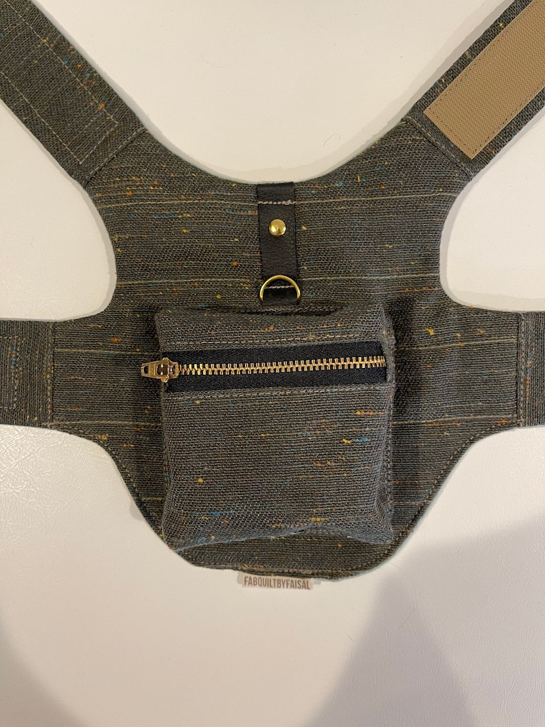 Pode incluir: Um arn&ecirc;s para c&atilde;o de lona verde escuro com um pequeno bolso com z&iacute;per e um anel de metal dourado. O arn&ecirc;s tem uma al&ccedil;a de couro marrom e um bot&atilde;o de metal dourado. O texto "FABQUILTBBYFAISAL" est&aacute; impresso na parte inferior da imagem.
