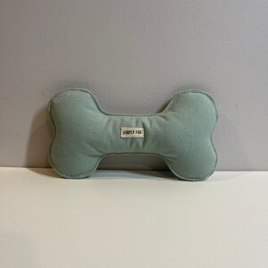 May include: A light green dog bone-shaped toy with a stitched outline. The toy has a small rectangular tag that reads "SIMPLY FAB". The toy is made of a soft, textured fabric.