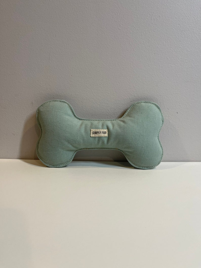 May include: A light green dog bone-shaped toy with a stitched outline. The toy has a small rectangular tag that reads "SIMPLY FAB". The toy is made of a soft, textured fabric.