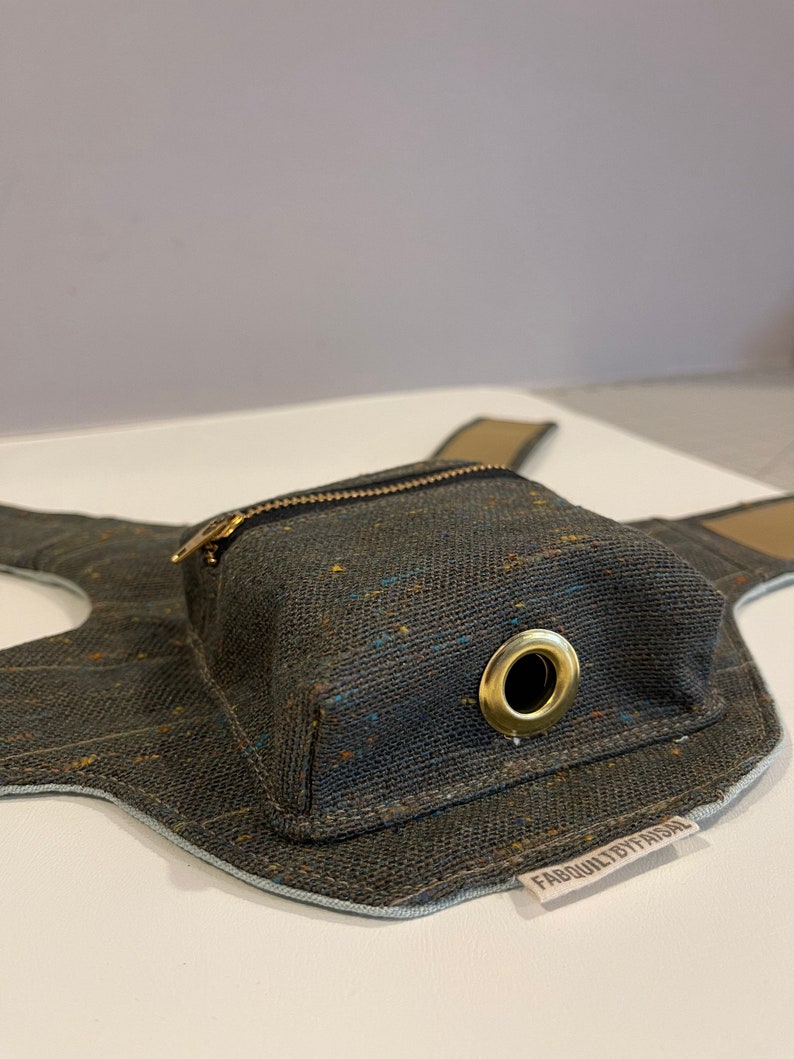 Pode incluir: Um arn&ecirc;s para c&atilde;o em lona cinzento escuro com um pequeno bolso com fecho de correr e um anel de metal dourado. O arn&ecirc;s tem uma guarni&ccedil;&atilde;o azul claro e um r&oacute;tulo castanho que diz "FABQUALITYBYFAISAL".