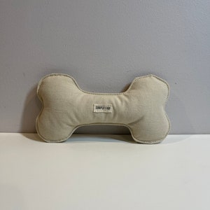 May include: A beige dog bone-shaped toy with a stitched outline. The toy has a rectangular label in the center with the text "SIMPLY FAB". The toy is made of a soft, textured fabric.