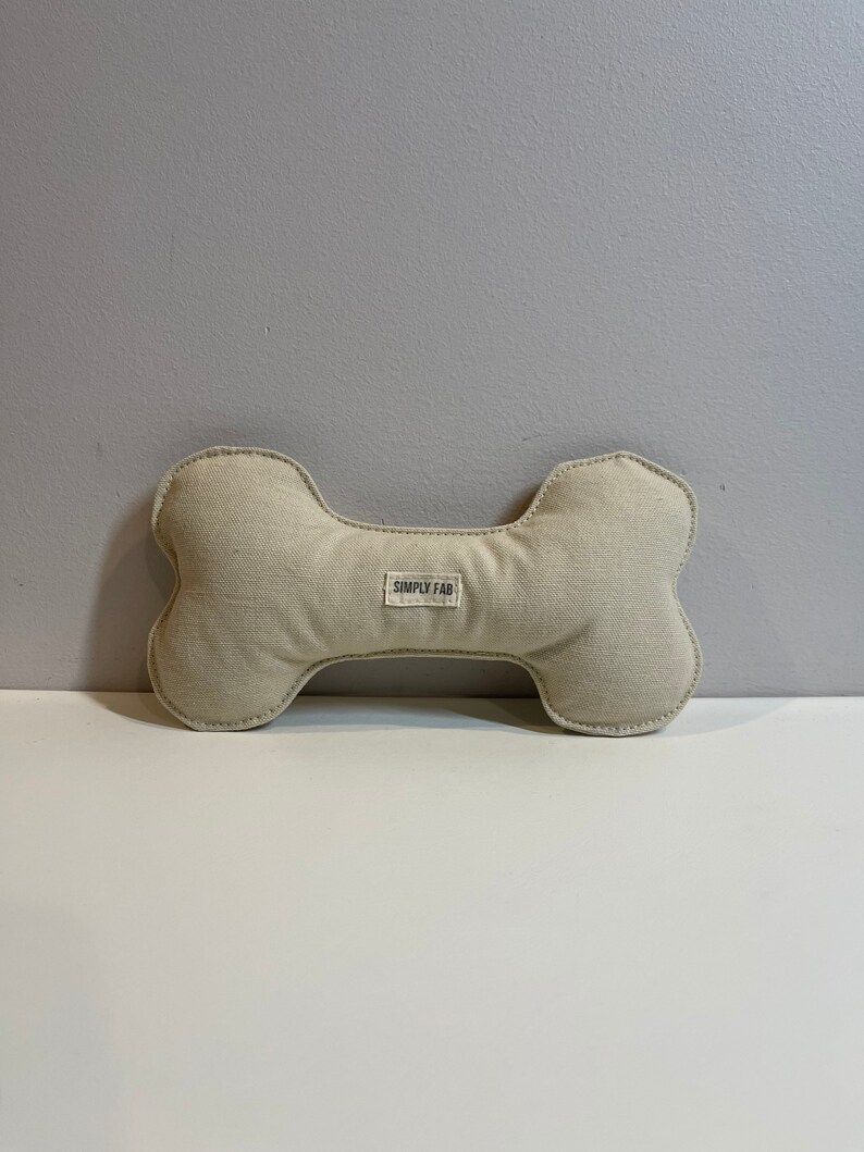 May include: A beige dog bone-shaped toy with a stitched outline. The toy has a rectangular label in the center with the text "SIMPLY FAB". The toy is made of a soft, textured fabric.