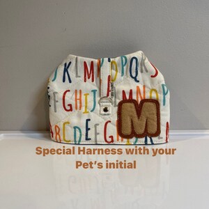 Op de afbeelding: Witte hondenharnas met een kleurrijke alfabetprint en een bruine viltletter M. "Special Harness with your Pet's initial" staat onder het harnas.