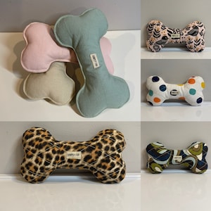 May include: A collection of dog bone-shaped toys in various colors and patterns. The toys include solid colors like pink, green, and tan, as well as patterned designs such as polka dots, floral, leopard print, and a retro swirl. Each toy has a "SIMPLY FAB" label.
