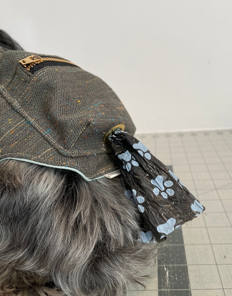 Pode incluir: Um porta-sacos para excrementos de c&atilde;es de lona cinza com um bolso com z&iacute;per e um design de pegada de pata azul. O porta-sacos est&aacute; preso ao colar de um c&atilde;o.
