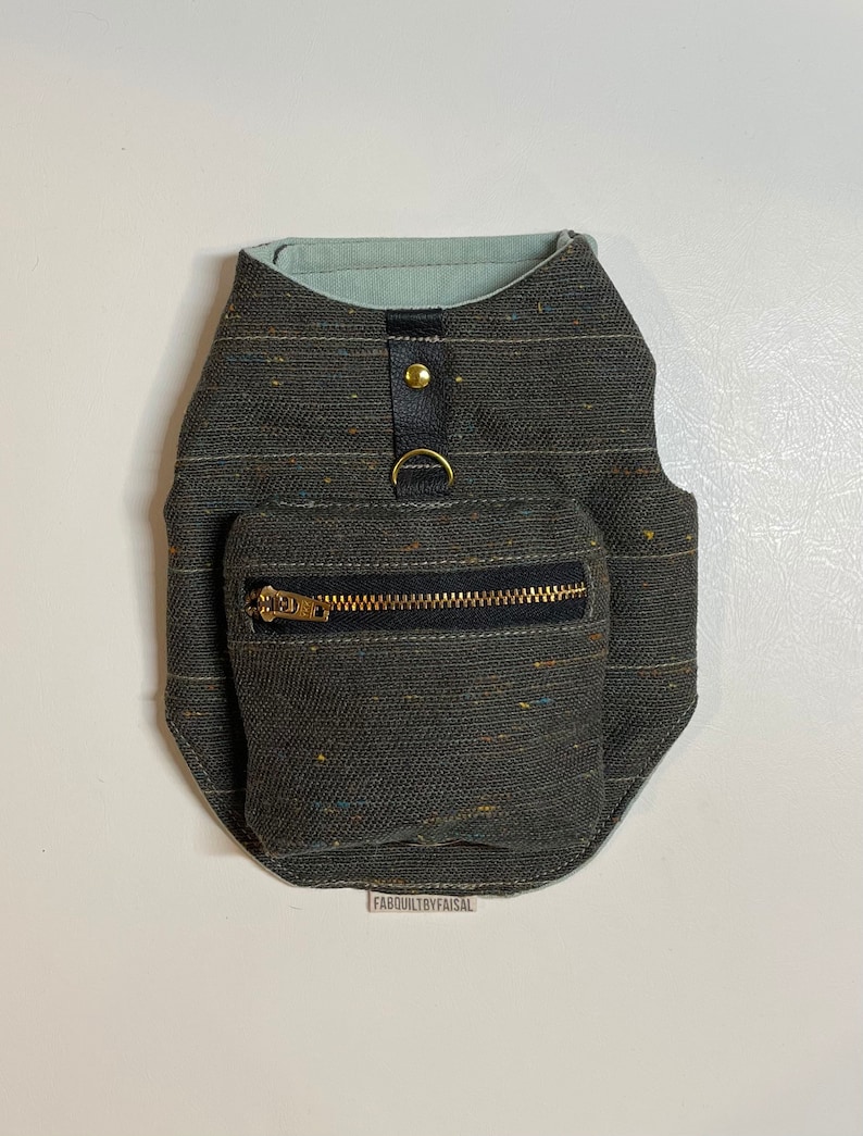 Pode incluir: Um arn&ecirc;s para c&atilde;o de cor verde escuro com um pequeno bolso com z&iacute;per e um anel de metal dourado. O arn&ecirc;s tem um forro de cor azul claro e um bot&atilde;o dourado.