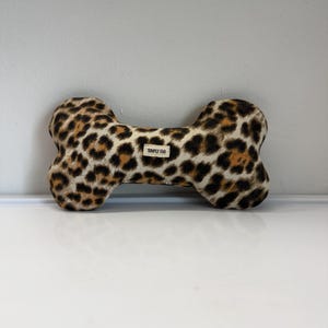 May include: A dog toy in the shape of a bone, featuring a leopard print pattern in shades of brown and black. The toy has a small tag that reads "SIMPLY FAB".