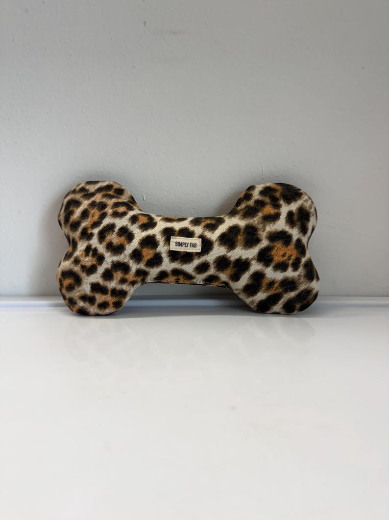May include: A dog toy in the shape of a bone, featuring a leopard print pattern in shades of brown and black. The toy has a small tag that reads "SIMPLY FAB".