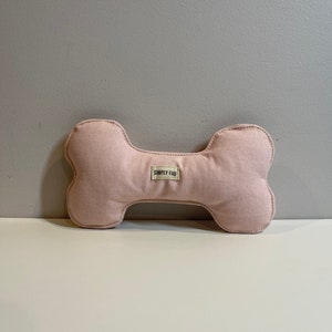 May include: A light pink dog bone-shaped toy made of canvas fabric. The toy has a stitched outline and a small label that reads "SIMPLY FAB". The toy is designed for pets.