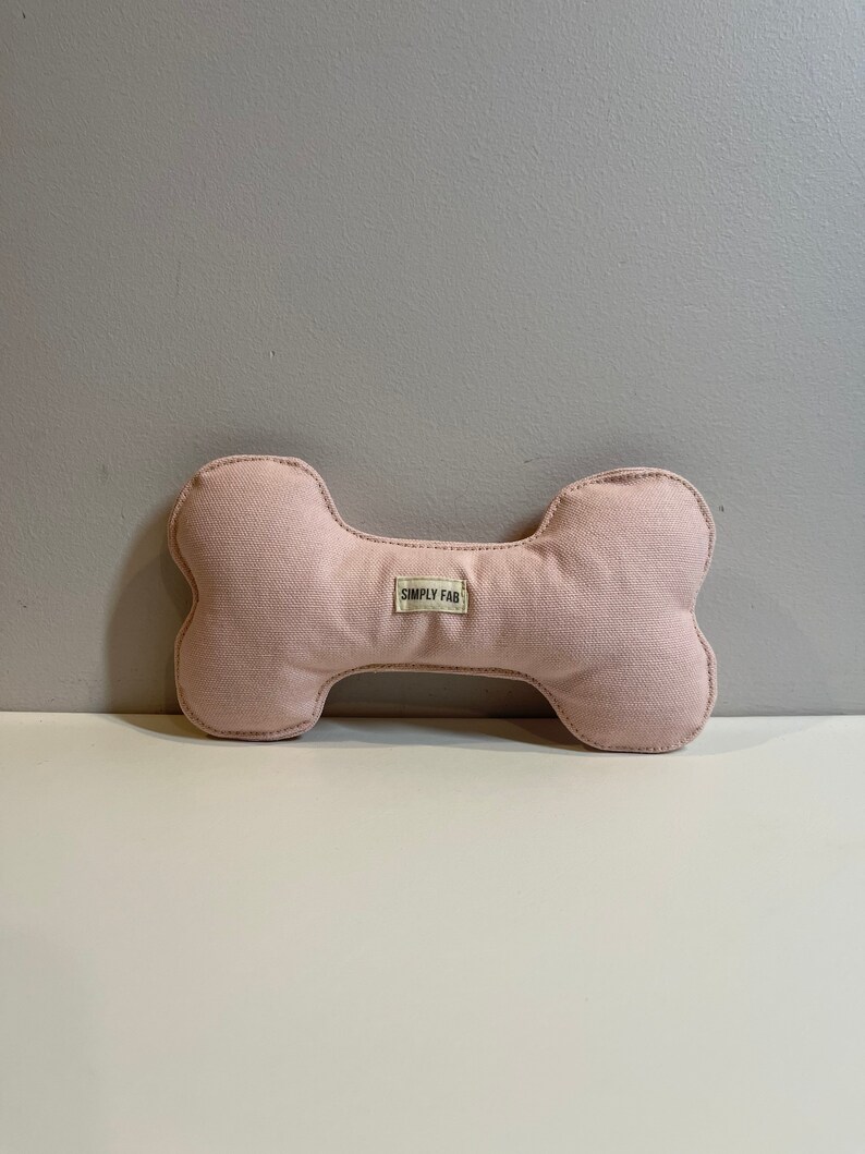May include: A light pink dog bone-shaped toy made of canvas fabric. The toy has a stitched outline and a small label that reads "SIMPLY FAB". The toy is designed for pets.